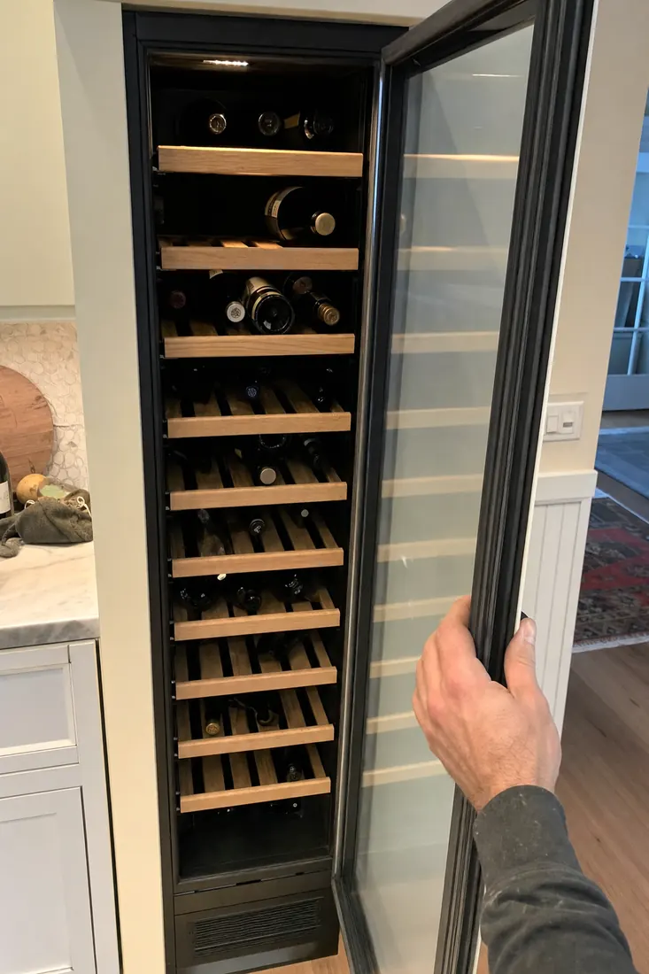 An open built-in wine column, bottles on wooden shelves, a hand at the door seal, bottles on their sides in frame - Buying & owning