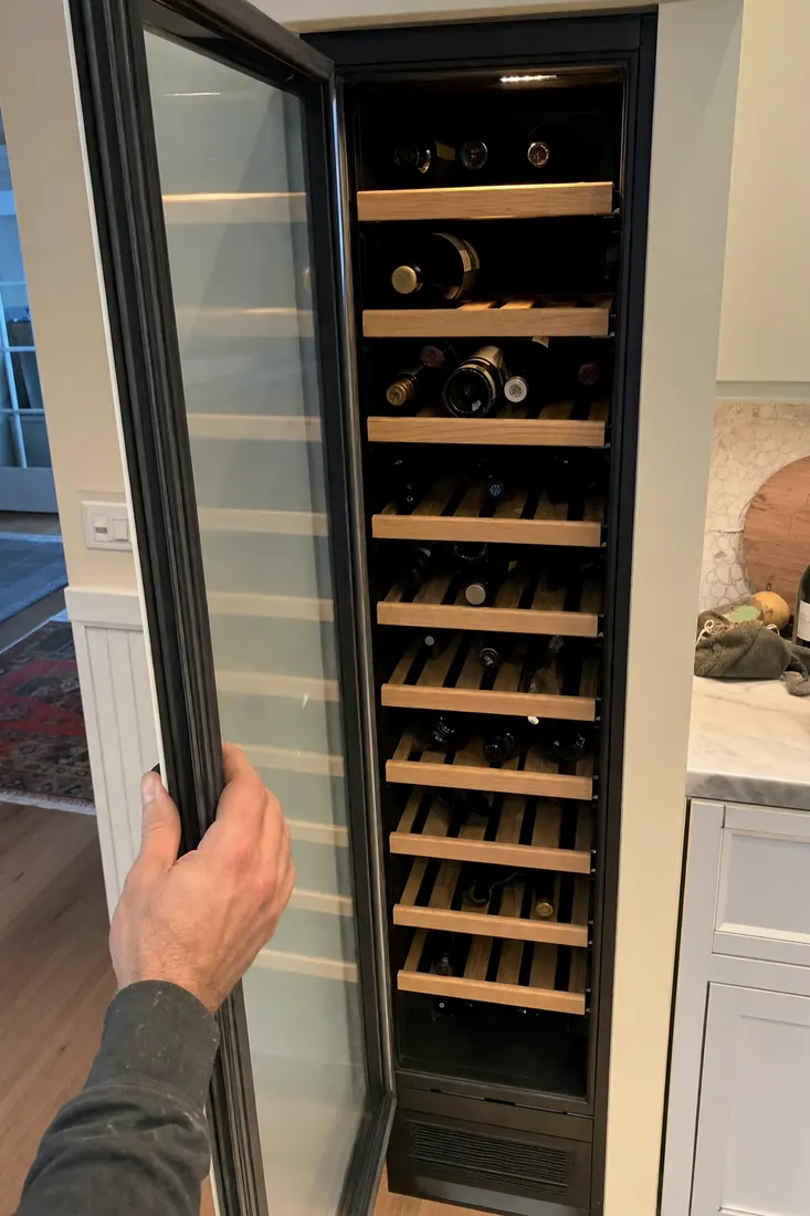 An open built-in wine column, bottles on wooden shelves, a hand at the door seal, the wooden shelves in frame - Repair or Replace a Built-In Appliance
