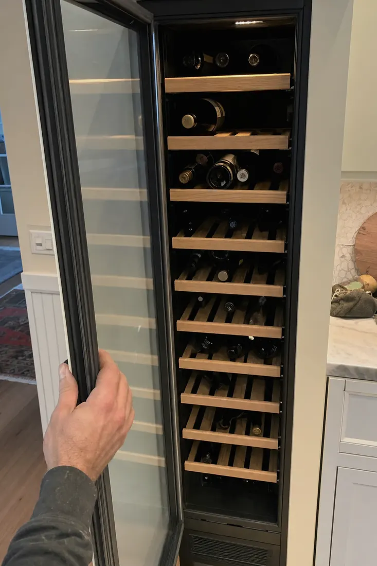 An open built-in wine column, bottles on wooden shelves, a hand at the door seal, the glass door in frame - What this costs, before we arrive