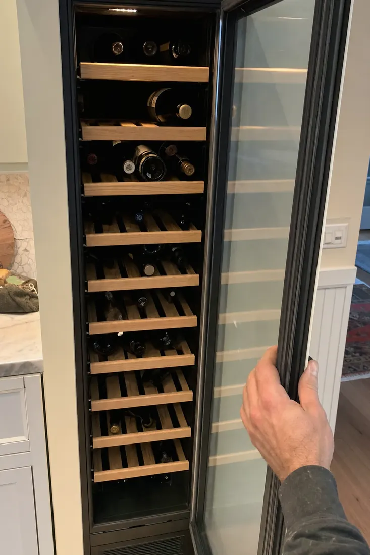 An open built-in wine column, bottles on wooden shelves, a hand at the door seal, the door gasket visible in the picture - Privacy Policy