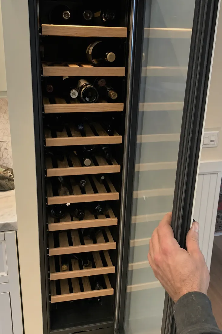 An open built-in wine column, bottles on wooden shelves, a hand at the door seal - Sub-Zero and luxury appliance repair in Neptune Beach