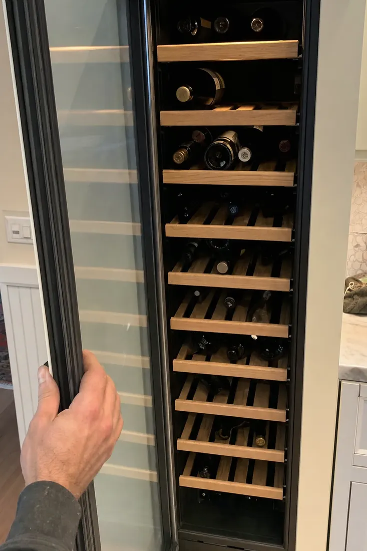 An open built-in wine column, bottles on wooden shelves, a hand at the door seal, the door gasket in frame - Sub-Zero and luxury appliance repair in Nocatee