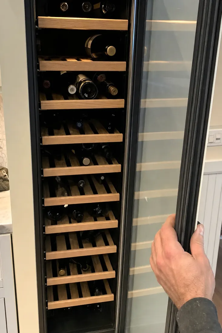 An open built-in wine column, bottles on wooden shelves, a hand at the door seal, bottles on their sides in frame - Sub-Zero and luxury appliance repair in Pablo Creek
