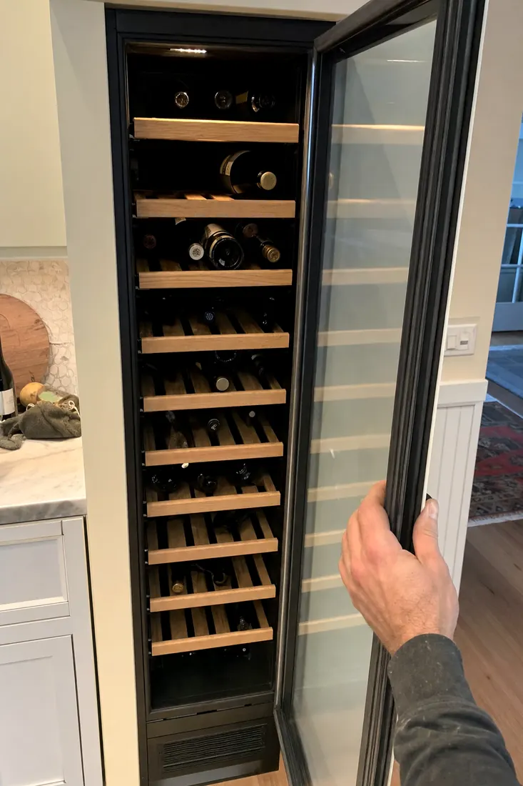 An open built-in wine column, bottles on wooden shelves, a hand at the door seal, the surrounding cabinetry in frame - Sub-Zero and luxury appliance repair in RiverTown