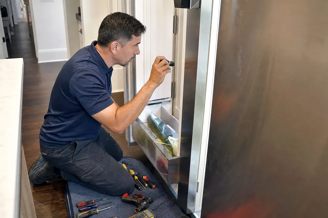 Technician checking inside an open built-in refrigerator with a flashlight, the kitchen floor in frame - True Residential repair in Jacksonville and Northeast