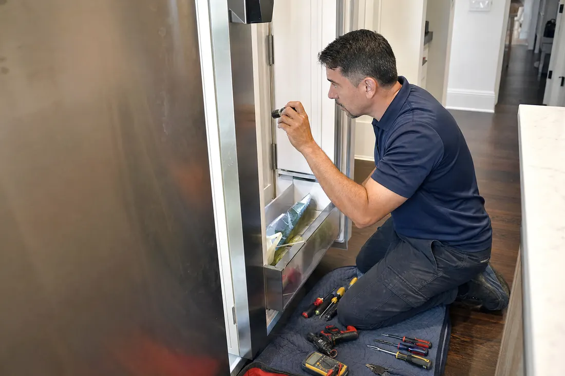 Technician checking inside an open built-in refrigerator with a flashlight, tools on a folded towel visible in the picture - Is It Worth Repairing a 20-Year-Old Sub-Zero? The Real