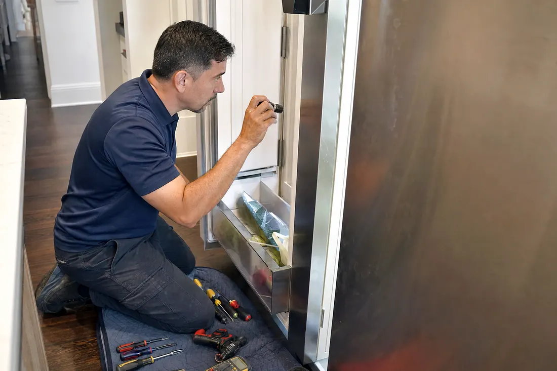 Technician checking inside an open built-in refrigerator with a flashlight, the kitchen floor visible in the picture - Water on the Floor Under Your Sub-Zero? Start