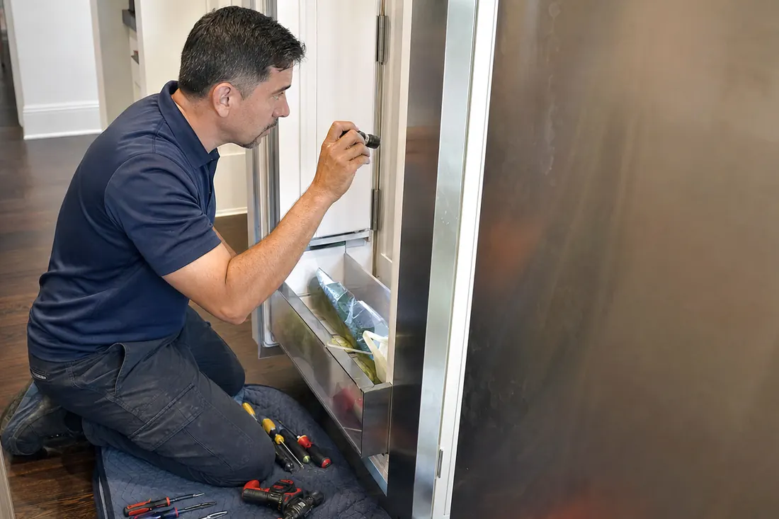 Technician checking inside an open built-in refrigerator with a flashlight, tools on a folded towel visible in the picture - Ice Maker and Undercounter Ice Machine Repair