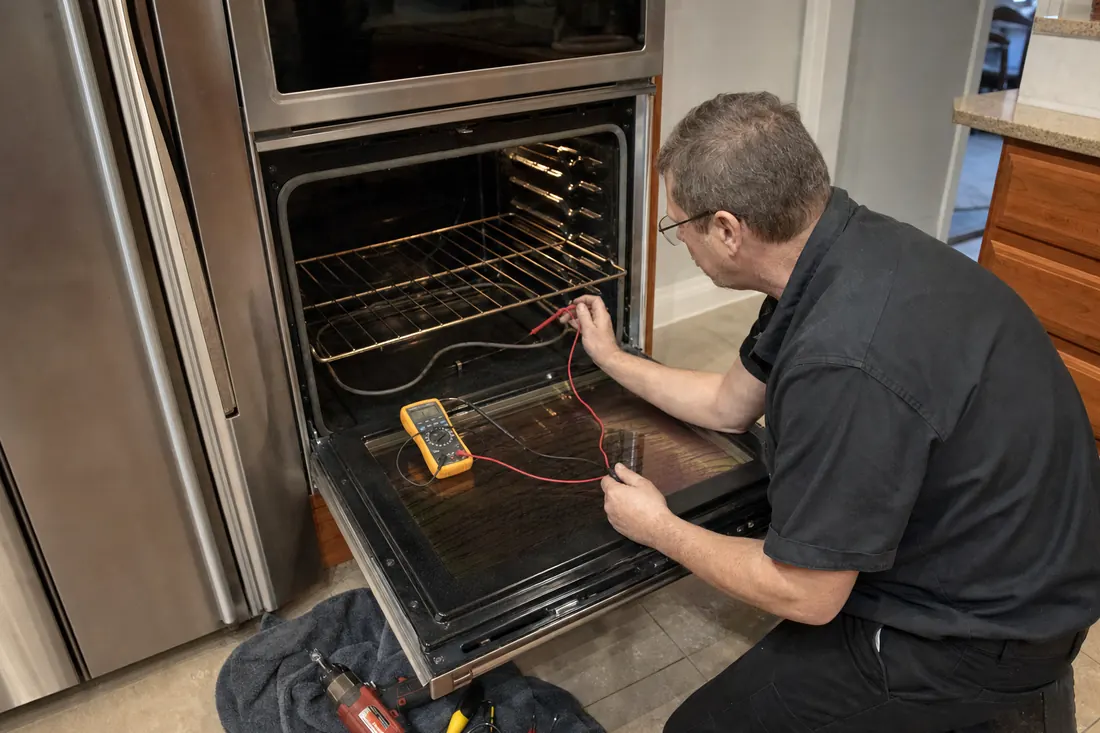 Technician testing the bake element of an open double wall oven with a multimeter, tools on a towel on the tile floor in frame - Built-In Coffee System Repair